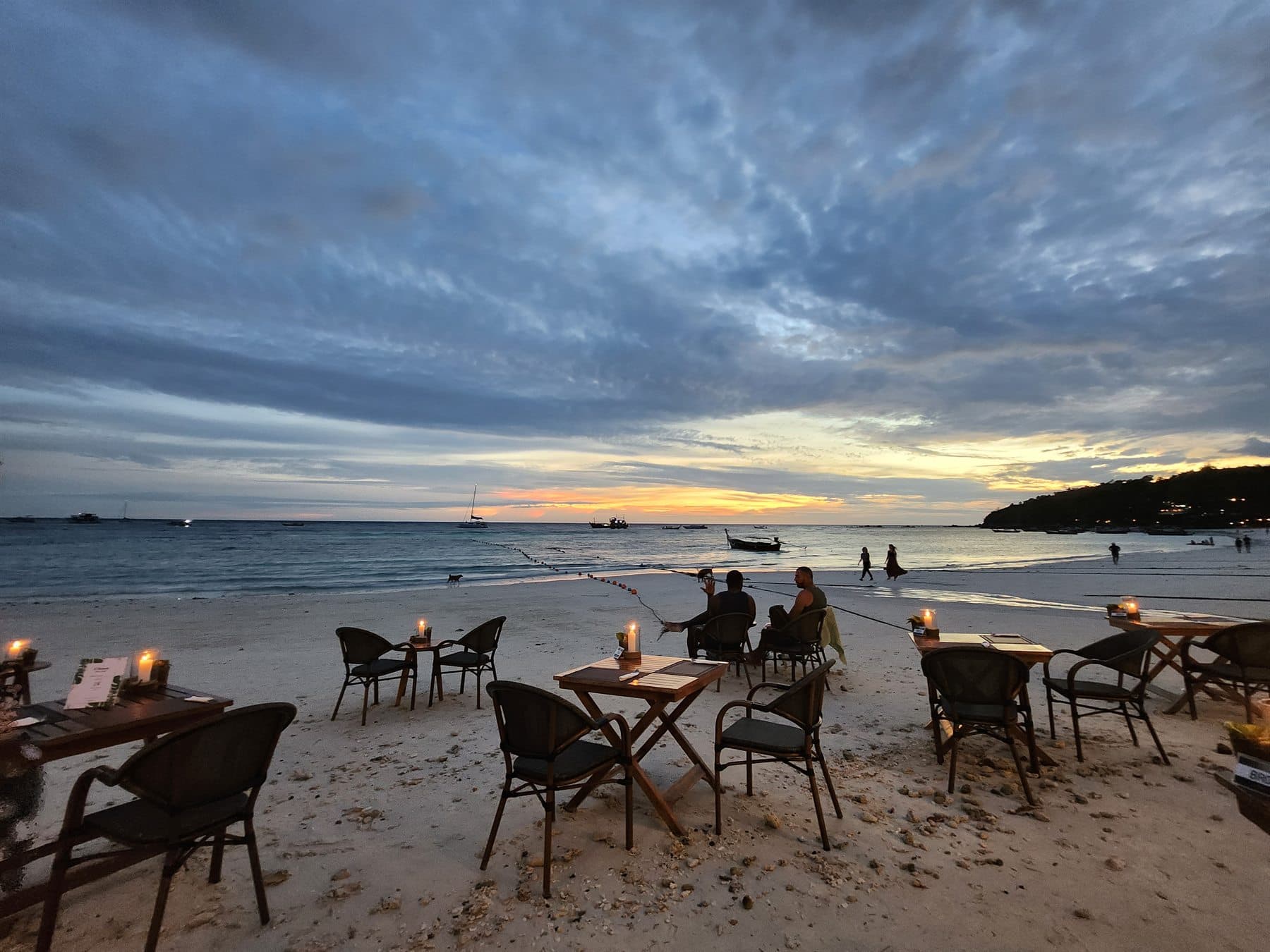Outdoor dining tables on a beach at sunset, with boats anchored offshore.